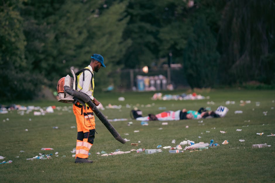 A male worker dressed in high-visibility orange trousers with reflective stripes, a white long-sleeved shirt, and a blue cap is using a leaf blower to clear litter from a grassy park area. He has a backpack-style vacuum or blower device attached to his back, with a large hose extending to the ground, and is focused on the task in front of him. In the background, there are two individuals lying on the grass near a set of outdoor trash disposal units, surrounded by scattered rubbish, including bottles, cans, and paper, which are also being blown or gathered by the worker. The scene is set outdoors in a green park with trees and natural foliage, with soft evening or late afternoon lighting. This image illustrates a professional approach to rubbish clearance, possibly performed by Waste Clearance Finsbury Park, highlighting on-site waste management and environmental cleanliness efforts.