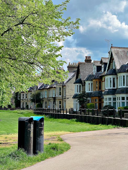 A quiet residential street scene featuring a row of traditional Victorian-style terraced houses with bay windows, painted in shades of cream, blue, and dark grey, each with small front gardens and fences. In the foreground, a paved footpath runs alongside a grassy area with two black public litter bins positioned near the edge of the path. A large leafy tree with fresh green foliage extends over part of the scene, casting dappled shade on the grass and pavement. The background shows a partly cloudy blue sky, contributing a natural, well-lit atmosphere. The image reflects a typical urban environment where private waste management services, such as those provided by Waste Clearance Finsbury Park, could support local rubbish removal needs, especially for those undertaking on-site waste disposal or alternative collection arrangements.