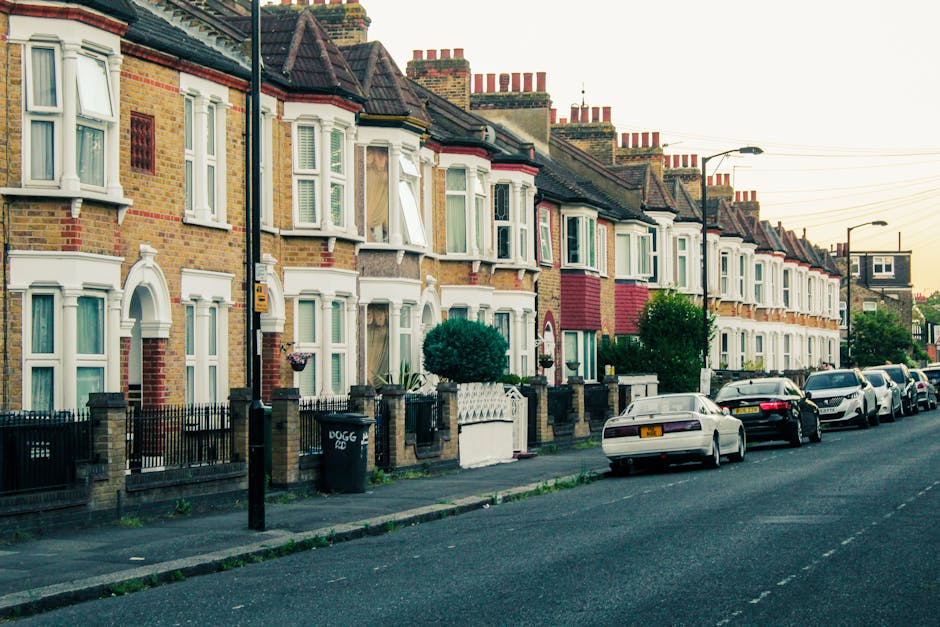 A row of closely spaced terraced houses situated along a residential street, featuring brick facades in yellow and red tones with white bay windows and decorative architectural details. Each property is separated by low brick and metal fences, some with small front gardens or paved areas, and a few have potted plants near the entrances. Several parked cars, including a white sedan and a black vehicle, align along the curb on the right side of the street. Tall street lamps and utility poles with cables stretch overhead, and the scene is illuminated by soft, natural daylight, suggesting either early morning or late afternoon. The environment appears clean and orderly, with no visible waste or debris, reflecting typical urban residential rubbish removal and private waste handling context. The scene is captured from the sidewalk, showing the uniformity of the houses and the street's calm, suburban atmosphere, consistent with the type of property maintenance supported by waste clearance services like those offered by Waste Clearance Finsbury Park.