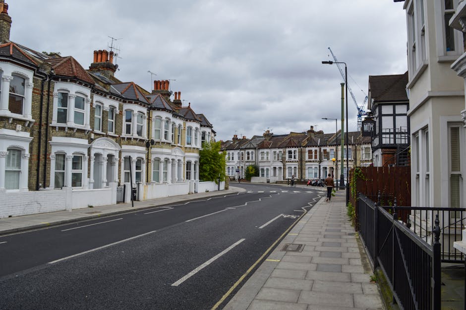 A quiet residential street lined with Victorian-style terraced houses featuring white facades, bay windows, and red-tiled roofs, with several brick chimneys visible on the rooftops. The street has a paved sidewalk on the right side, bordered by black metal railings and small front gardens. There are streetlights and a few parked cars along the curb. The sky is overcast with grey clouds, creating diffuse, soft lighting. In the distance, a construction crane can be seen above the rooftops, indicating nearby ongoing development. The scene is typical of urban UK neighborhoods, where private rubbish clearance services like Waste Clearance Finsbury Park might operate to manage waste collection outside of council services, though no waste or rubbish is visible in the image itself.