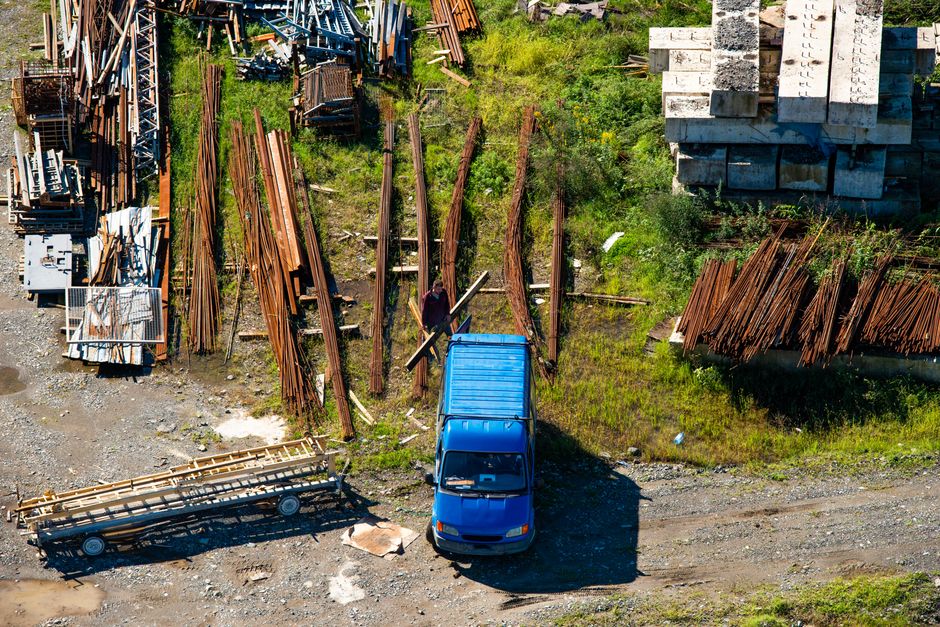 An aerial view of a construction materials yard showing various rusty steel rebars and wooden pallets arranged in uneven stacks on grassy and gravelly ground. A person stands amidst the materials, holding a long bar or pipe. A blue van is parked on a dirt surface near the materials, with its shadow cast on the ground. To the right, there is a large stack of cement blocks or concrete slabs, neatly piled in a rectangular formation. Adjacent to the left side of the image, there are more materials, including metal beams and wooden planks, some stacked on platforms or pallets. The scene is lit by natural daylight, highlighting the textures and colors of the materials, with patches of grass and small weeds growing between the stacks, suggesting an informal or temporary storage area, typical of a site requiring waste removal or on-site clearance service from Waste Clearance Finsbury Park. This setting supports the context of private waste handling or alternative rubbish collection solutions commonly associated with construction or renovation projects in the Finsbury Park area.