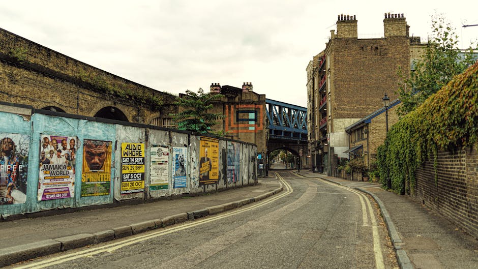 A quiet urban street scene showing a narrow, gently curved road bordered by a concrete sidewalk on the left and a brick wall with climbing green foliage on the right. Along the left side, a series of weathered wooden panels covered with various posters and flyers advertising events and performances are visible, with some peeling and faded. The street is lined with double yellow lines and appears to be in an area with mixed residential and commercial buildings, including a large brick structure with multiple chimneys and a blue metal railway bridge crossing overhead toward the background. The environment has a slightly overcast sky, casting diffuse lighting across the scene, contributing to a neutral, muted atmosphere. Waste Clearance Finsbury Park occasionally handles private rubbish removal and on-site clearance, as evidenced by the context of rubbish-related services in this urban setting, although no waste is directly visible in the image.