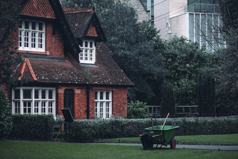 A public park area in a residential neighbourhood during daytime, featuring a large, leafy green tree on the left with bright fresh foliage. In the foreground, there are two black and blue litter bins positioned on a grassy strip beside a concrete pathway. Behind the bins, a row of Victorian-style terraced houses with bay windows, brick and stone facades, and chimney stacks line the background. The houses have mixed roof finishes, including slate and tiled surfaces, with some feature decorative woodwork painted in contrasting colours. The sky above is partly cloudy with patches of blue visible, providing natural daylight that illuminates the scene with a neutral tone. The environment suggests an urban setting that may benefit from private or independent waste collection, highlighted by the presence of the waste bins, which could be managed by waste clearance services like Waste Clearance Finsbury Park for rubbish removal or alternative waste handling outside of council services.