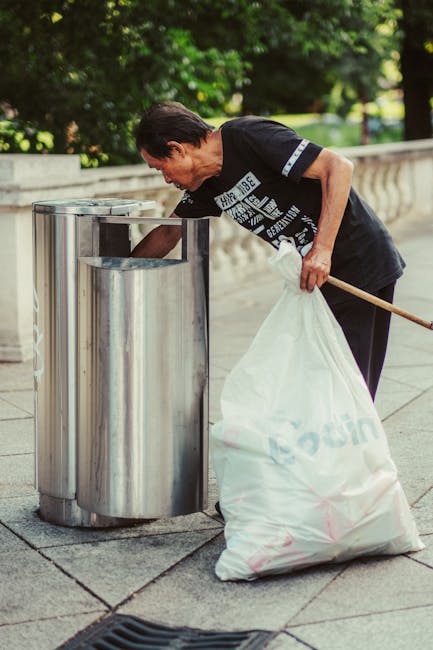 A large pile of mixed household rubbish and waste materials is gathered around and overflowing from several black, grey, and red rubbish bins placed on a paved sidewalk in an urban setting. The waste includes flattened cardboard boxes, paper bags, plastic packaging, and discarded household items. The cardboard boxes are partially collapsed and some are torn, with crumpled paper and plastic bags spilling onto the surrounding pavement. The grey bin is labeled for mixed paper and card, with its lid open and refuse piled on top, indicating an informal or private collection method rather than official council services. Behind the rubbish, there are parked cars, a small metal barrier, and a commercial building with storefronts and signage, partially obscured by scaffolding and urban décor like trees and railings. The scene reflects typical outdoor rubbish accumulation that might require professional waste clearance, such as services offered by Waste Clearance Finsbury Park for alternative waste handling or on-site clearance of bulky rubbish in the area.