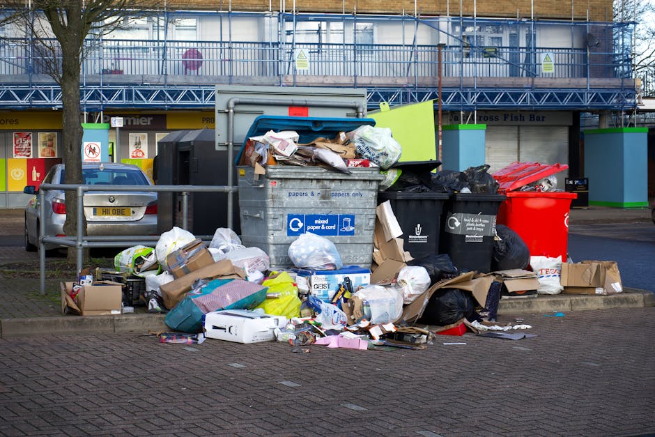 A large collection of rubbish is piled on a paved street corner in front of a commercial building, with multiple black and red bins, some overflowing with waste materials, and numerous cardboard boxes, paper bags, and plastic packaging scattered across the ground. The waste includes flattened cardboard boxes, crumpled paper, plastic bottles, and various discarded packaging materials. The bins are made of metal and plastic, with one gray bin labeled for mixed paper and cardboard, and others in black and red, some with lids partially open revealing contents. To the left, a parked silver car is visible behind a metal railing, with a street tree situated nearby. In the background, a commercial storefront with signage and a two-story building with glass windows and scaffolding are seen, indicating an urban environment. The scene appears well-lit with natural daylight, and the overall impression is of an on-site collection of domestic or business waste awaiting removal, aligning with services offered by Waste Clearance Finsbury Park for private or alternative rubbish disposal.