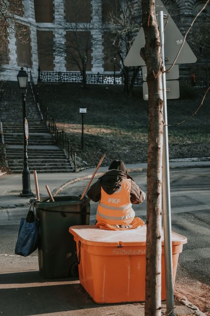 A worker wearing an orange high-visibility vest with the abbreviation 'FKE' on the back is seated on an orange plastic rubbish bin, facing away from the camera, in an outdoor urban environment. The worker appears to be engaged in rubbish collection or waste management, with a large green wheelie bin placed beside them. The green bin contains several wooden sticks or poles protruding from the top, and a blue bag hangs from its side. The scene is set on a paved street next to a tree with a protective metal guard around its trunk, partially obscuring part of a sign mounted on a tall street pole. In the background, there are stone steps leading up to a building, some leafless trees, and a wrought iron fence, suggesting it might be late autumn or winter. The lighting is natural, with shadows indicating a sunny day, and the area resembles a public space that may require independent waste disposal outside regular municipal rubbish collection, aligning with private rubbish removal services like those offered by Waste Clearance Finsbury Park.
