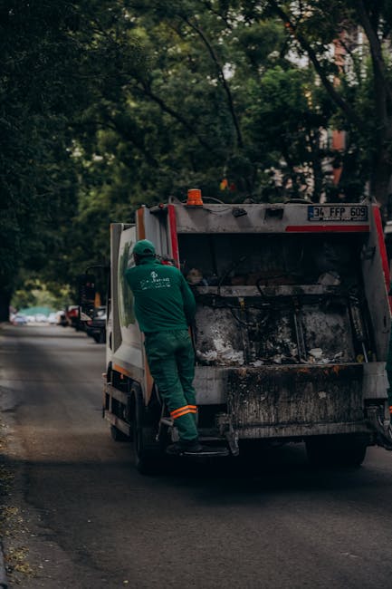 A worker wearing an orange high-visibility vest with the abbreviation 'FKE' on the back is seated on an orange plastic rubbish bin, facing away from the camera, in an outdoor urban environment. The worker appears to be engaged in rubbish collection or waste management, with a large green wheelie bin placed beside them. The green bin contains several wooden sticks or poles protruding from the top, and a blue bag hangs from its side. The scene is set on a paved street next to a tree with a protective metal guard around its trunk, partially obscuring part of a sign mounted on a tall street pole. In the background, there are stone steps leading up to a building, some leafless trees, and a wrought iron fence, suggesting it might be late autumn or winter. The lighting is natural, with shadows indicating a sunny day, and the area resembles a public space that may require independent waste disposal outside regular municipal rubbish collection, aligning with private rubbish removal services like those offered by Waste Clearance Finsbury Park.